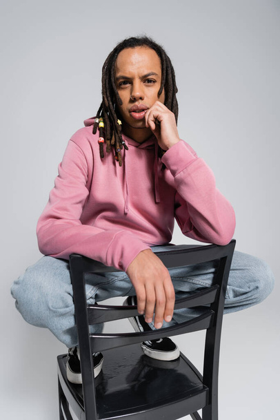 pierced multiracial man looking at camera while sitting in shoes on chair isolated on grey  - Foto, afbeelding