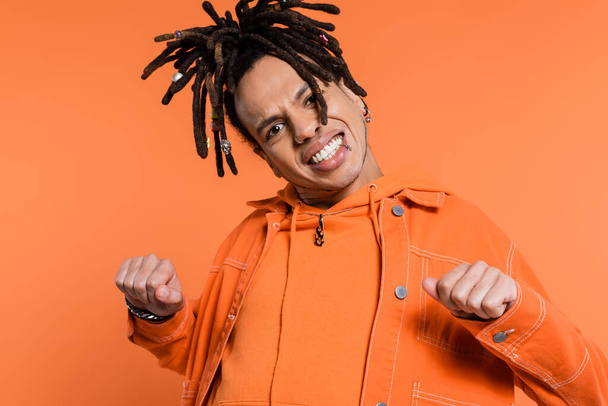 excited and multiracial man with dreadlocks looking at camera while standing with clenched fists isolated on coral background  - Фото, изображение