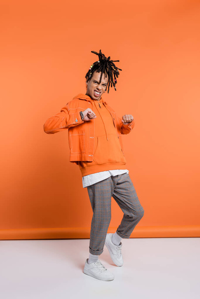 full length of excited and multiracial man with dreadlocks standing with clenched fists on coral background  - Photo, Image