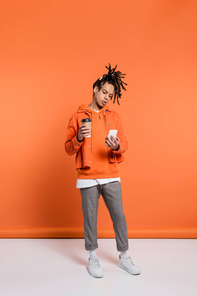 full length of pierced and multiracial man with dreadlocks holding paper cup and using smartphone on coral background  - Zdjęcie, obraz