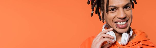 cheerful and multiracial man with wireless headphones looking at camera isolated on coral background, banner  - Foto, afbeelding