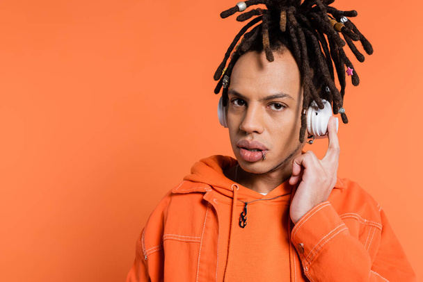 pierced and multiracial man listening music in wireless headphones while looking at camera on coral background  - Valokuva, kuva
