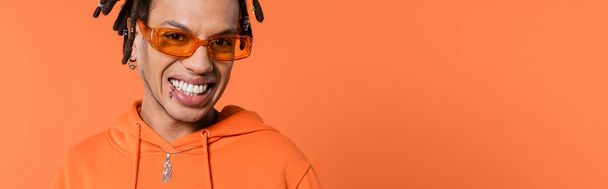 happy multiracial man with dreadlocks wearing stylish sunglasses isolated on coral background, banner  - 写真・画像