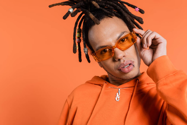 multiracial man with dreadlocks adjusting trendy sunglasses isolated on coral background  - Foto, imagen