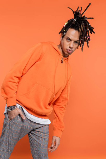 pierced multiracial man with dreadlocks standing with hand in pocket isolated on coral background  - Photo, Image