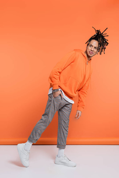 full length of pleased multiracial man with dreadlocks standing with hand in pocket on coral background  - Фото, зображення