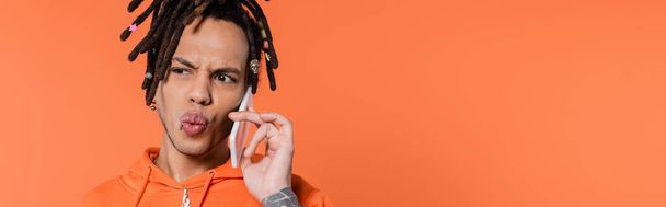 multiracial man with dreadlocks pouting lips and talking on cellphone isolated on coral background, banner - Photo, image