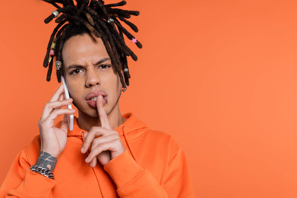 multiracial man with dreadlocks holding finger near lips and talking on cellphone isolated on coral  - 写真・画像