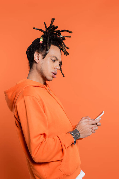 vista lateral del hombre multirracial tatuado con dreadlocks mensajes de texto en el teléfono inteligente aislado en coral  - Foto, Imagen