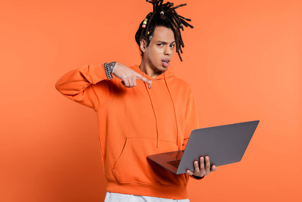 verward multiraciale man met dreadlocks en tatoeage wijzen naar laptop op koraal achtergrond  - Foto, afbeelding