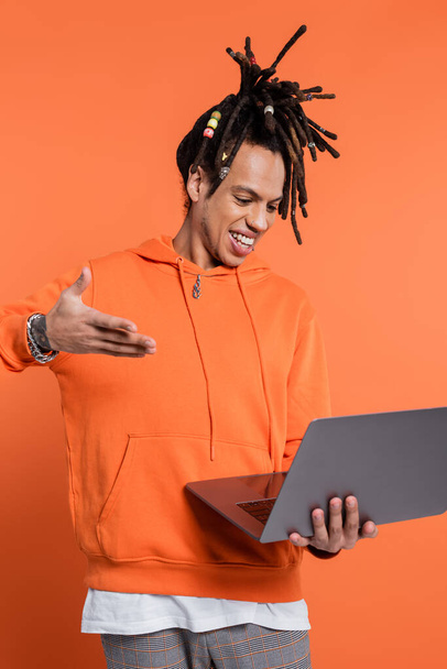 happy multiracial man with dreadlocks and tattoo pointing at laptop on coral background  - Foto, Bild