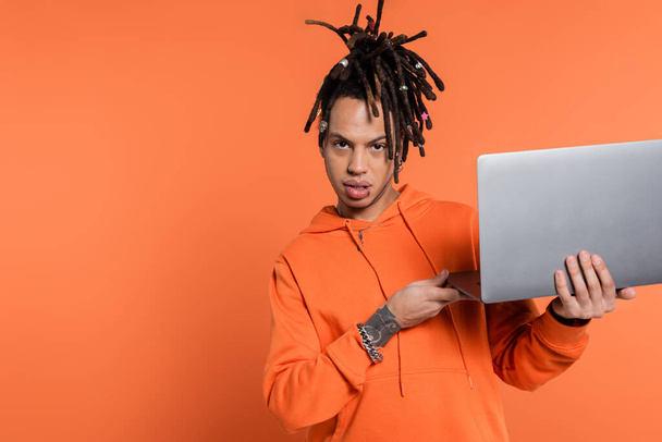 multiracial freelancer with dreadlocks and tattoo holding laptop and looking at camera on coral background  - Photo, image