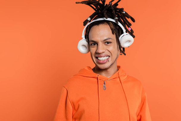 funny multiracial man with dreadlocks and wireless headphones isolated on coral  - Foto, Imagen