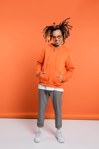 full length of happy multiracial man in hoodie and stylish sunglasses posing with hands in pockets on coral background - Foto, afbeelding