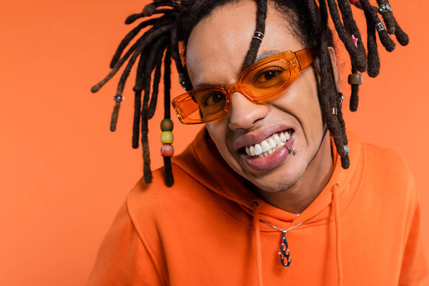 portrait of satisfied multiracial man in hoodie and stylish sunglasses isolated on coral  - Fotoğraf, Görsel
