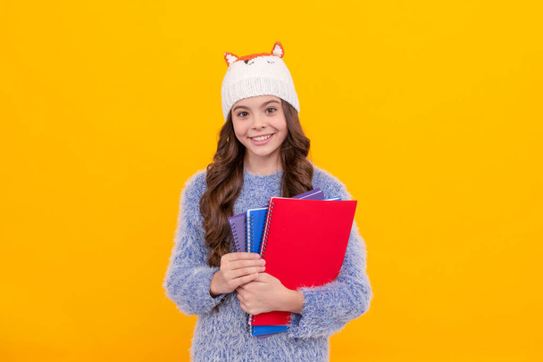 happy kid with curly hair in hat. back to school. teen girl in knitwear on yellow background. portrait of child holding workbook. express positive emotion. winter fashion. - Photo, image