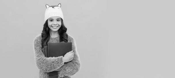 Winterschule. Pullover und Hut halten Arbeitsmappe auf gelbem Hintergrund. Porträt einer Schülerin, Kopfzeile eines Atelierbanners. Schulkindgesicht, Kopierraum - Foto, Bild