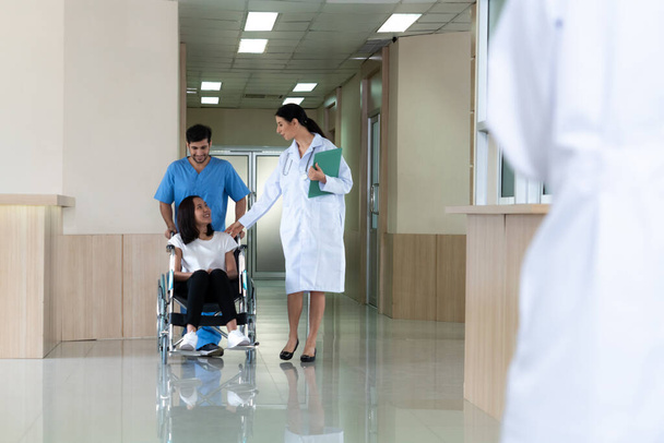 医師と男性看護師は、滅菌病院の廊下に沿って車椅子の女性患者を輸送します。病院構想における障害者のための医療・介護. - 写真・画像