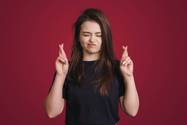 Hopeful brunette woman crossing fingers for good luck being dressed in a black t-shirt closing eyes isolated over red background. Female student making wish - Foto, Bild