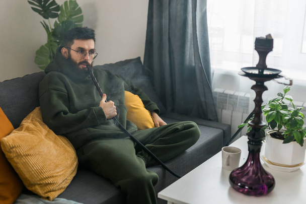 Bearded millennial or gen z man smoking hookah while relaxing on sofa at home - chill time and resting - Foto, imagen