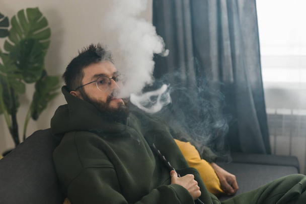 Γενειάδα χιλιετή ή gen z άνθρωπος κάπνισμα hookah, ενώ χαλαρώνοντας στον καναπέ στο σπίτι - chill time and resting - Φωτογραφία, εικόνα