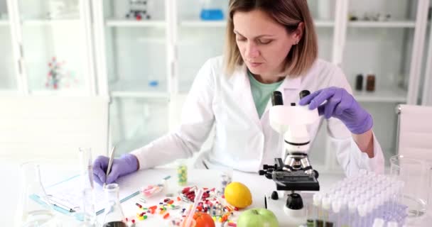Female scientist works on microscope and analyzes fruits and vegetables and writes notes in reports. Harmful food factors and natural toxins - Séquence, vidéo