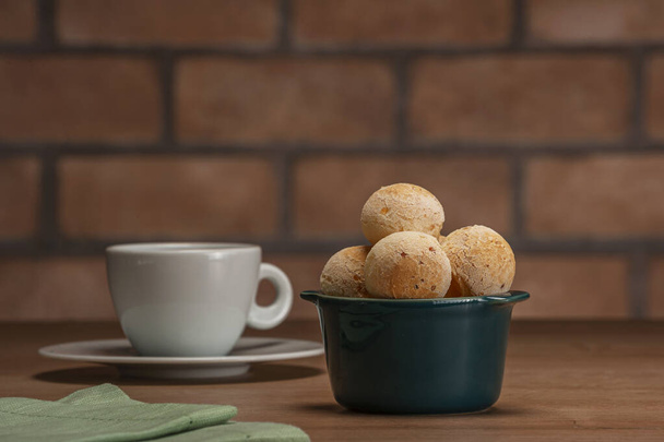 Cheese breads in a green ramekin with a cup of coffee on wooden table and bricks wall background. - Photo, image