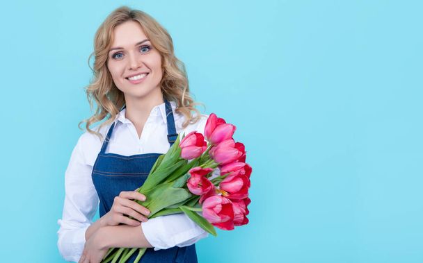 cheerful young woman in apron with spring tulip flowers on blue background. - Photo, Image