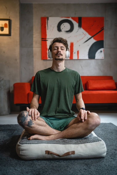 ένας άνδρας ενήλικας καυκάσιος άντρας στο πάτωμα χρησιμοποιεί ακουστικά για online καθοδηγούμενο διαλογισμό ασκώντας mindfulness yoga με τα μάτια κλειστά στο σπίτι πραγματικοί άνθρωποι αυτο-φροντίδα εκδήλωση έννοια αντιγραφή χώρου - Φωτογραφία, εικόνα