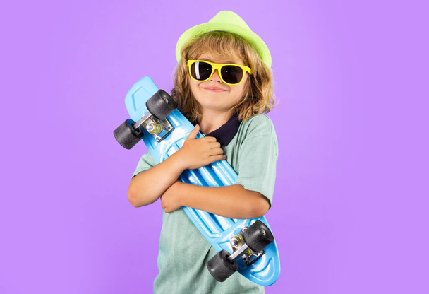 Παιδί της μόδας με καλοκαιρινό πουκάμισο κομψό καπέλο και γυαλιά ηλίου. Χαριτωμένο παιδί με skateboard στο χρώμα ισοβαθμισμένο φόντο. Αστείο παιδί αγόρι, κομψό σκέιτερ κρατώντας skateboard στο στούντιο - Φωτογραφία, εικόνα
