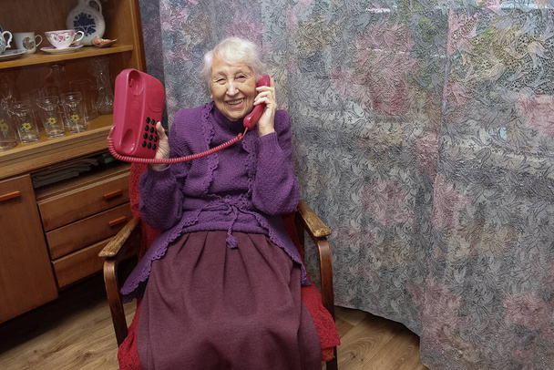 Mujer mayor feliz usando teléfono rojo fijo de estilo retro en casa - Foto, imagen