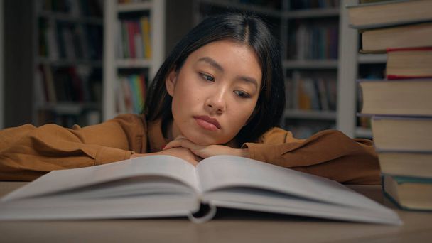 Βαριέται λυπημένη κουρασμένη τεμπέλα νεαρή Κορεάτισσα να διαβάζει βαρετό βιβλίο ξαπλωμένη στο γραφείο της στη βιβλιοθήκη του πανεπιστημίου χωρίς κίνητρα φοιτήτρια που προετοιμάζεται για τις εξετάσεις του κολλεγίου υπερφορτώνεται με μελέτη αντιπαθητικό θέμα - Φωτογραφία, εικόνα