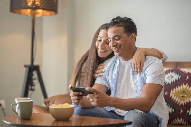 Ευτυχισμένο νεαρό ευτυχισμένο ζευγάρι που χρησιμοποιεί εφαρμογές κοινωνικής δικτύωσης smartphone στο σπίτι, χαμογελαστοί σύζυγοι πελάτες της χιλιετίας που μιλούν για δέσιμο βλέποντας αστείο βίντεο κοιτάζοντας το κινητό τηλέφωνο χαλαρώνοντας στον καναπέ - Φωτογραφία, εικόνα