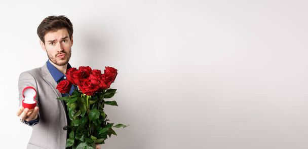 Handsome boyfriend in suit asking to marry him, standing with red bouquet of roses and engagement ring, looking pleading at camera, standing over white background. - Foto, Imagem