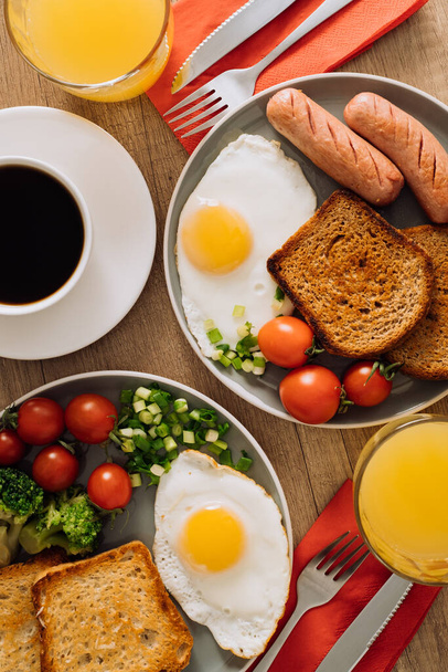 Επίπεδη lay αγγλικό πρωινό με φλιτζάνι μαύρο καφέ και χυμό πορτοκαλιού, λουκάνικο σχάρας και τοστ ολικής αλέσεως με τηγανητό αυγό και ντοματίνια - Φωτογραφία, εικόνα