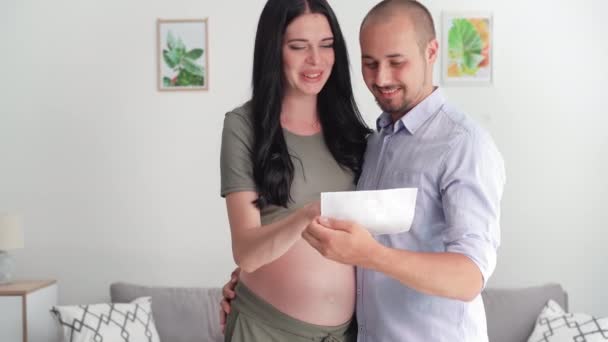 raskaus, muotokuva onnellisista vanhemmista, jotka pitävät ultraäänikuvan käsissä, hymyilevät ja katsovat kameraa - Materiaali, video