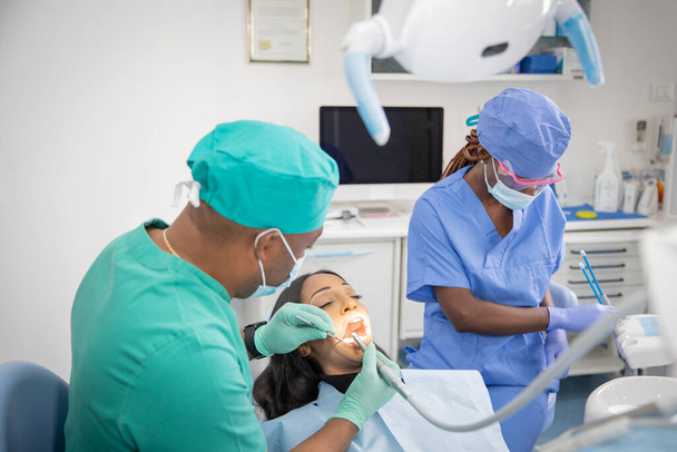 Afrykański dentysta sprawdza stan zdrowia pacjenta podczas gdy jego asystent przygotowuje narzędzia pracy.. - Zdjęcie, obraz