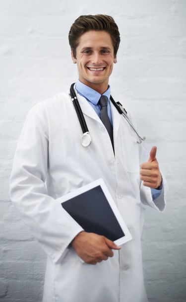 Sağlıklı bir tıbbi onay. Tablet taşıyan yakışıklı genç bir doktorun portresi ve baş parmak işareti. - Fotoğraf, Görsel