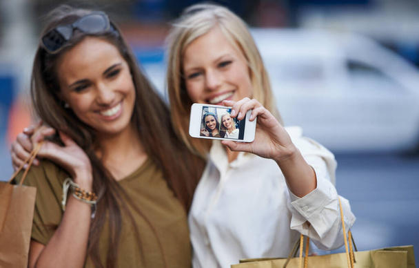 Φτιάχνω αναμνήσεις μεταξύ φίλων. Δύο νεαρές γυναίκες να κρατάνε τσάντες για ψώνια και να φωτογραφίζουν τον εαυτό τους σε ένα κινητό. - Φωτογραφία, εικόνα