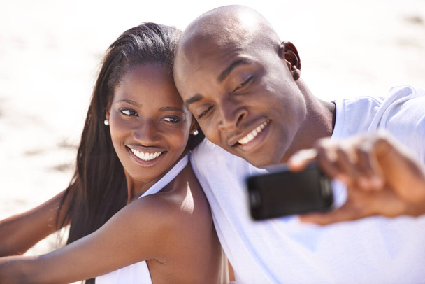 Lächeln Sie für mich... Ein glückliches junges afrikanisches Paar lächelt, während es einen Schnappschuss am Strand macht - Foto, Bild