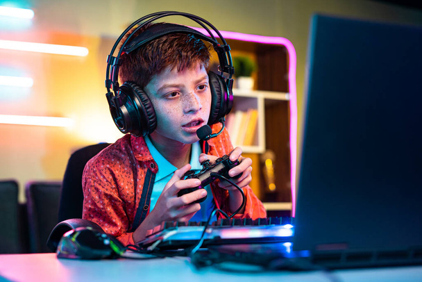 Teenager kid playing online video gameon laptop by talking on headphones at home - concept of pro gamer, tournament and live streaming - Photo, Image