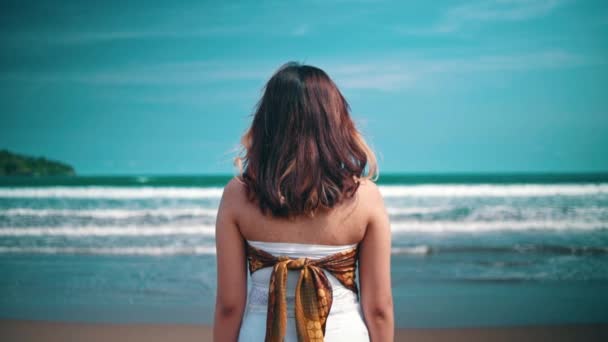 An Asian woman in a white dress is standing on the beach while looking at the beautiful view of the blue sea and the waves during the day - Metraje, vídeo