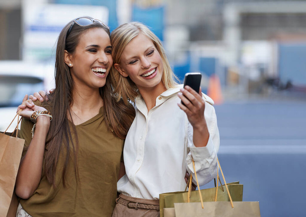 Harika görünüyoruz. Alışveriş torbaları tutan ve cep telefonuyla kendi portresini çeken iki genç kadın. - Fotoğraf, Görsel