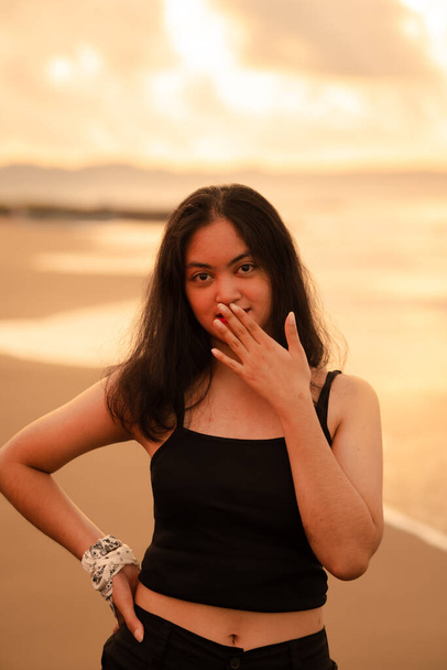 une femme asiatique avec des vêtements noirs et de longs cheveux noirs debout sur le sable de la plage avec une expression plate pendant les vacances le matin - Photo, image
