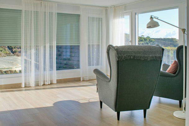 Acogedor interior de la casa de luz con grandes ventanas, persianas, cortinas y cómodo sillón. - Foto, imagen