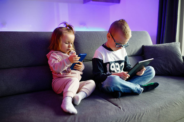 Даня и его младшая сестра Эмилия играют с мобильными телефонами и планшетами. Дети постоянно сидят на телефонах и планшетах. - Фото, изображение