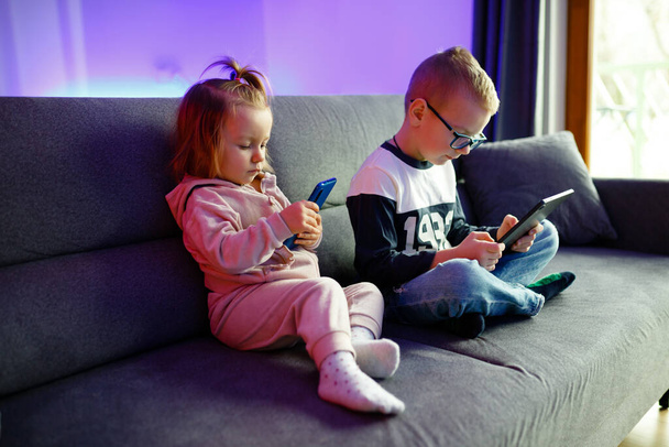 Danya and his younger sister Emilia play with mobile phones and tablets. Children are constantly sitting on phones and tablets. - Foto, Imagen