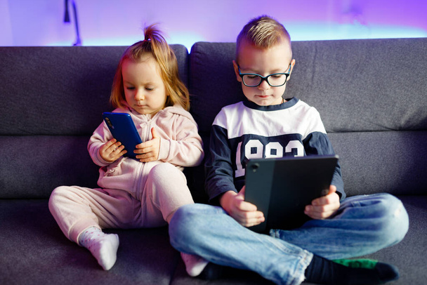 Danya y su hermana menor Emilia juegan con teléfonos móviles y tabletas. Los niños están constantemente sentados en teléfonos y tabletas. - Foto, Imagen