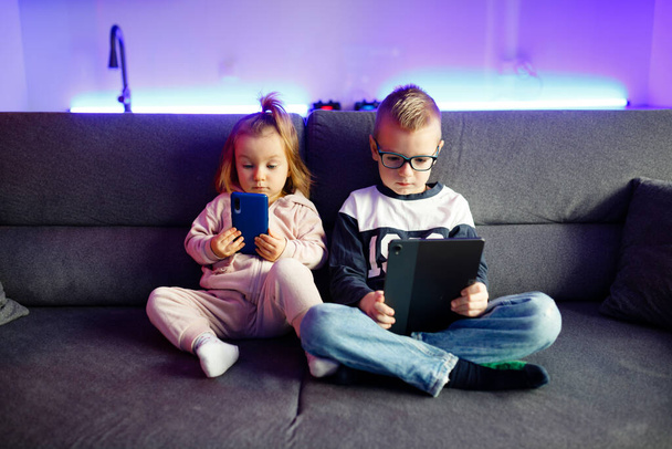 Danya ja hänen pikkusiskonsa Emilia leikkivät matkapuhelimilla ja tableteilla. Lapset istuvat jatkuvasti puhelimissa ja tableteissa. - Valokuva, kuva