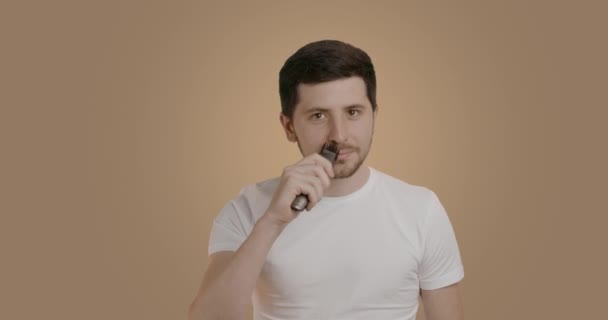 Dark-haired man with smooth skin arranges his beard with the help of an electric shaver. Young man shaving his beard with a razor on a nude studio background. Grooming concept for men - Imágenes, Vídeo